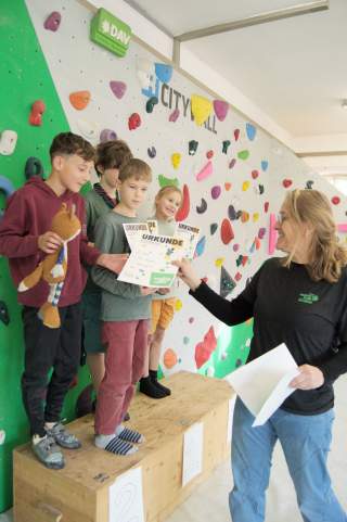 Vier Kinder stehen auf einer Siegertreppe vor einer Kletterwand und erhalten von einer Frau Urkunden überreicht. Ein Kind hält ein Stofftier.