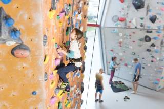 Ein Kind klettert an einer bunten Kletterwand in einer Kletterhalle, im Hintergrund stehen drei Erwachsene vor einer weiteren Kletterwand.