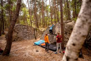 Der Lärchenwald von Fontainebleau in dem zwei große Sandsteine zu sehen sind. An dem rechten bouldert eine Frau in blauer Jacke. Zwei weitere Sportlerinnen schauen ihr gespannt zu. Zur Sicherheit sind am Fuß des Felsen einige Crashpads ausgelegt.
