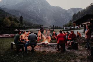 Um ein Lagerfeuer sitzen 15 Jugendliche auf Holzbänken. Etwas abseits stehen drei Schlauchbote. Am rechten Bildrand sind eine Holzhütte und eine Wäscheleine zu sehen. Der Hintergrund wird von einem Berg ausgefüllt.