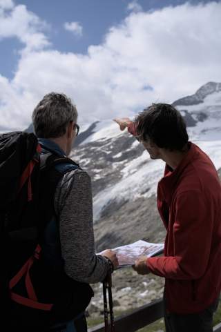 Zwei Wanderer mit Rucksack und Karte stehen vor schneebedeckten Bergen, während einer der beiden auf die Landschaft zeigt.