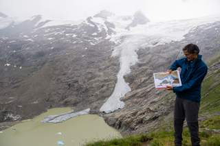 Mann in blauer Jacke steht auf einer Bergwiese und hält eine Karte, im Hintergrund ist ein Gletscher mit Schmelzwassersee zu sehen.