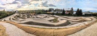 Panoramaaufnahme einer kurvigen und hügeligen Fahrradstrecke im Freien, umgeben von Bäumen und Wohngebäuden im Hintergrund.