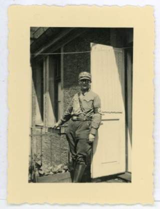 Schwarzweißfoto eines Mannes in NSDAP-Uniform mit Mütze und Hakenkreuzarmbinde, der vor einer offenen Haustür steht und in die Kamera blickt.