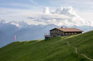 Hütte auf einer grünen Bergwiese mit Alpenpanorama und Wolken im Hintergrund.