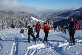 Vier Menschen mit Skiern und Stöcken stehen lachend im verschneiten Gebirge, weitere Personen folgen im Hintergrund, umgeben von schneebedeckten Tannen und Bergen.