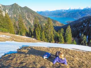 Eine Frau sitzt auf einem teils noch verschneitem Berghang im Gras und genießt den Frühlingstag mit Blick auf einen See.