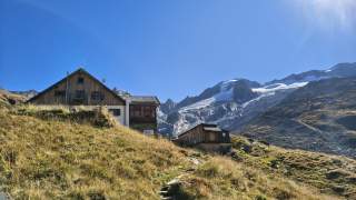 Hütte und Nebengebäude auf Bergwiese vor vergletscherten Bergen.