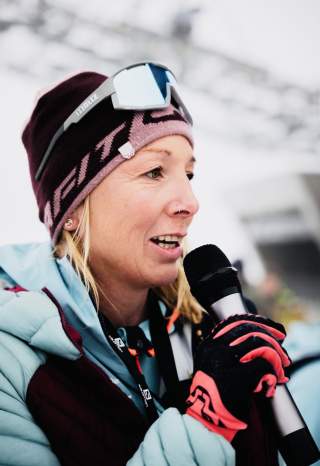 Frau in Winterkleidung mit Mütze, Skibrille und Handschuhen spricht in ein Mikrofon.