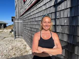 Frau in schwarzem Tanktop mit verschränkten Armen lehnt an einer Holzschindelwand und lächelt in die Sonne.