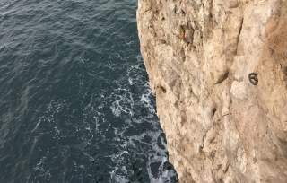 Blick auf eine steile Felswand über dem Meer, an der ein Kletterhaken befestigt ist.