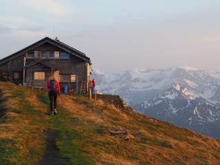 Eine Person wandert zu einer Berghütte, im Hintergrund sind schneebedeckte Berge im Abendlicht zu sehen.