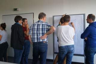 Eine Gruppe von Menschen steht vor einem Whiteboard, auf dem Notizen gemacht werden, und diskutiert. Einige blicken nachdenklich, während zwei Personen aktiv schreiben.