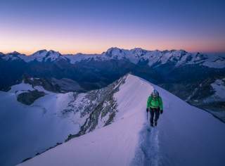 Bergsteiger mit Hochtourenausrüstung auf schneebedecktem Gipfelgrat, im Hintergrund Gipfelpanorama vor Abend- oder Morgenrot
