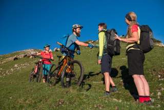 Ein Mann und eine Frau kommen je ein Mountainbike schiebend auf einem Grashang von oben, ein Mann und eine Frau kommen ihnen zu Fuß von unten entgegen, die beiden Männer geben sich freundlich die Hand. Im Hintergrund ist eine Schafherden zu sehen.