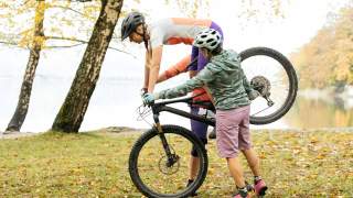 Mountainbike-Fahrerin übt nur auf dem Vorderrad zu stehen und wird auf beiden Seiten von 2 Personen gehalten, im Hintergrund ein See und Herbstbäume