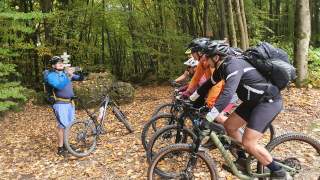 4 Personen stehen nebeneinander in einer Reihe auf dem Mountainbike im Wald, eine weitere macht ein Foto mit dem Smartphone von der Gruppe