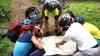 7 Personen mit Fahrradhelmen sitzen in der Hocke auf einem Waldweg um eine Wanderkarte, 1 Person deutet auf die Karte, im Hintergrund liegen Mountainbikes