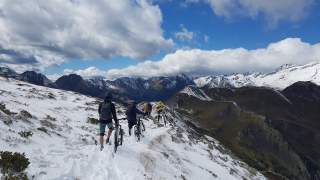 4 Personen mit Fahrradhelmen und Rucksäcken schieben hintereinander ihre Mountainbikes auf einem Bergrücken durch einige Zentimeter Neuschnee, im Hintergrund Berggipfel