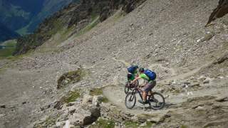 2 Personen fahren hintereinander mit dem Mountainbike bergab durch Schotter