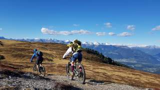 2 Personen auf dem Mountainbike fahren auf einem schmalen Schotterweg einen breiten Bergrücken entlang, die Wiesen sind braun, im Hintergrund schneebedecktes Gipfelpanorama