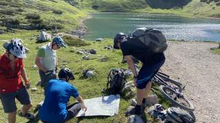 3 Personen mit Fahradhelmen stehen auf einer Grasfläche neben einem Schotterweg um eine Wanderkarte, 1 Person hockt vor der Karte und zeigt darauf, im Hintergrund ein Bergsee, daneben ein liegendes Mountainbike und Rucksäcke