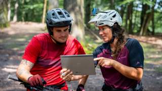 Ein Mountainbiker auf dem Fahrrad mit Helm stehend und eine Mountainbikerin mit Helm und Tablet sehen sich gemeinsam etwas auf dem Tablet an