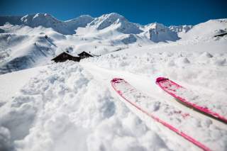 pinkfarbene, mit Schnee bedeckte Skispitzen im Vordergrund, im Hintergrund eine Hütte und Berggipfel schneebedeckt