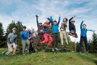 9 Mitglieder des Lehrteams Mountainbike springen auf einer Graskuppe in die Luft, im Hintergrund 2 liegen 2 Mountainbikes