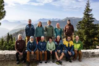 7 Mitglieder des Lehrteams Naturschutz sitzen in erster Reihe auf einer Steinmauer, 5 weitere stehen in 2. Reihe dahinter, im Hintergrund Ausblick auf Berge im blauen Dunst