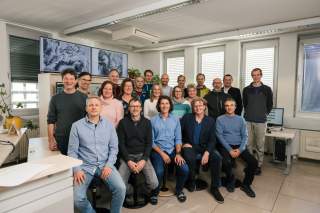 Gruppenfoto von etwa 20 Personen in einem modernen Büro mit mehreren Computermonitoren und Pflanzen, einige sitzen, andere stehen, alle lächeln in die Kamera.