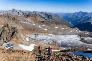 Zwei Wanderer mit Helmen und Rucksäcken stehen auf einem felsigen Bergpfad und blicken auf eine weite Gebirgslandschaft mit Gletscherresten und einer Seilbahnstation. Der Himmel ist klar und blau.