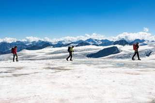 Drei Bergsteigende mit Ausrüstung wandern über einen schneebedeckten Gletscher vor einer Kulisse von schroffen Bergen und blauem Himmel.