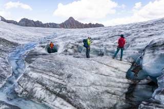 drei Menschen stehen auf einem Gletscher, ein Schmelzwasserbach fließt die Eisfläche hinab, eine der Personen steht vor einer Gletschermühle
