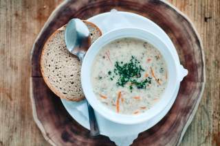 Auf einem Tablett steht eine Schüssel mit Gerstensuppe, mit Schnittlauch bestreut. Daneben ein Löffel und eine Scheibe Mischbrot.