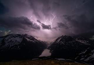 Über den noch verschneiten Alpengipfeln und einem Stausee blitzt es.