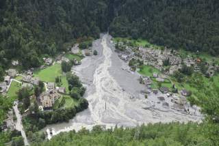 Luftaufnahme eines Dorfes in einem Tal, durch das ein breiter Schlammlawinenstrom verläuft und Teile der Häuser und Landschaft verwüstet hat.