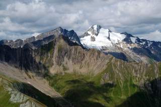 Berglandschaft mit schroffen Felsen, grünen Wiesen und schneebedecktem Gipfel unter bewölktem Himmel.