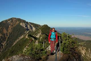 Frau mit Rucksack und Sonnenbrille wandert auf einem schmalen Bergpfad mit Sicherungsseilen, im Hintergrund Berge und weiter Landschaftsblick unter blauem Himmel.