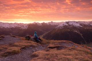 Person sitzt auf Bank am Berg in rötlichem Sonnenuntergang