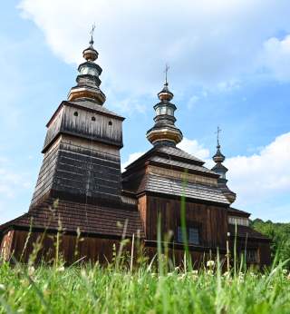 Alte Holzkirche mit mehreren Zwiebeltürmen, aufgenommen aus der Froschperspektive, im Vordergrund grünes Gras, im Hintergrund blauer Himmel mit Wolken.