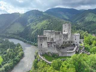 Alte Burgruine auf einem grünen Hügel oberhalb eines Flusses, umgeben von dichten Wäldern und Bergen.