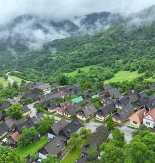 Luftaufnahme eines kleinen Dorfes mit vielen Häusern, umgeben von grünen Hügeln und Wäldern, im Hintergrund nebelverhangene Berge.
