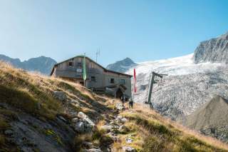 Berghütte auf einer Wiese vor schneebedeckten Bergen, mit Wanderern und Seilbahnstütze im Hintergrund.