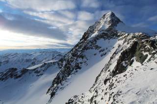 Verschneiter, felsiger Berggipfel unter einem bewölkten Himmel mit Ausblick auf eine Bergkette im Hintergrund.