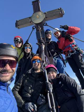Eine Gruppe von sieben Bergsteigerinnen und Bergsteigern posiert lächelnd an einem Gipfelkreuz vor blauem Himmel.