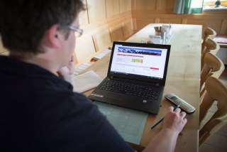 Person sitzt an einem Holztisch und arbeitet an einem Laptop, daneben liegen ein Telefon und Papiere.