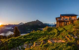 Eine Berghütte steht auf einer grünen Wiese mit Blick auf schneebedeckte Berge im Hintergrund, während die Sonne am Horizont untergeht.