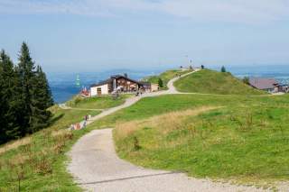 Ein geschotterter Weg auf einem weiten Bergrücken führt zu einer Berghütte, im Hintergrund erkennt man flaches Land.