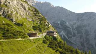 Eine Alpenvereinshütte vor Gebirgskulisse, im Vordergrund grüne, karge Wiesen.